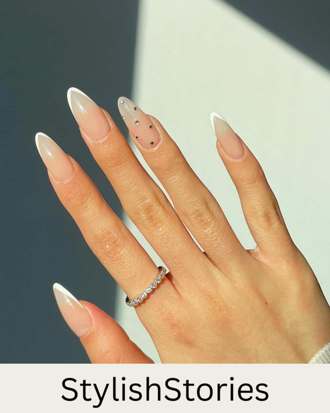 A hand with long almond nails painted with pointed white tips and a crystal accent nail