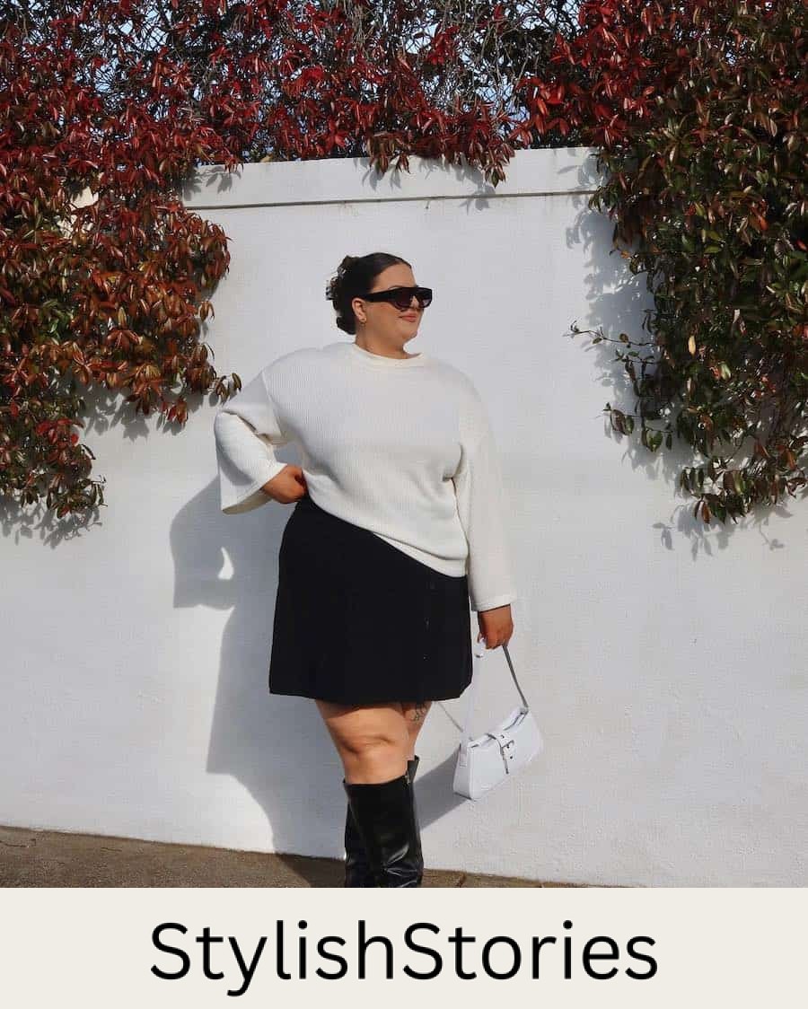 A woman wearing a black mini skirt, a white sweater, and tall black boots with a white handbag