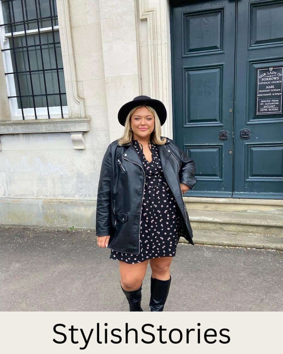 A woman wearing a black floral mini dress with tall black leather boots, a black leather jacket, and a black fedora