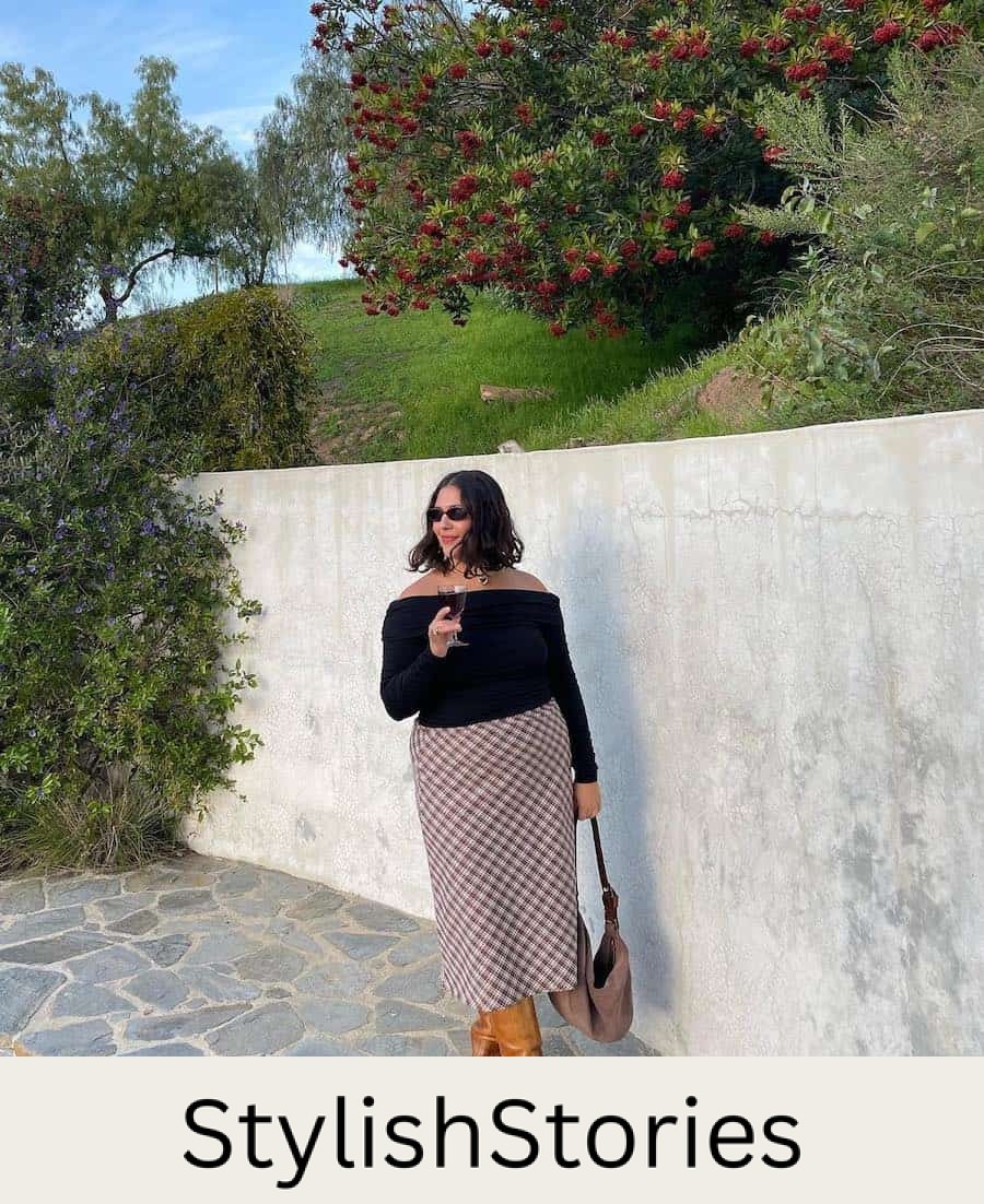 A woman wearing a white and red plaid midi skirt with an off-the-shoulder black sweater, tall brown boots, and a brown hobo bag