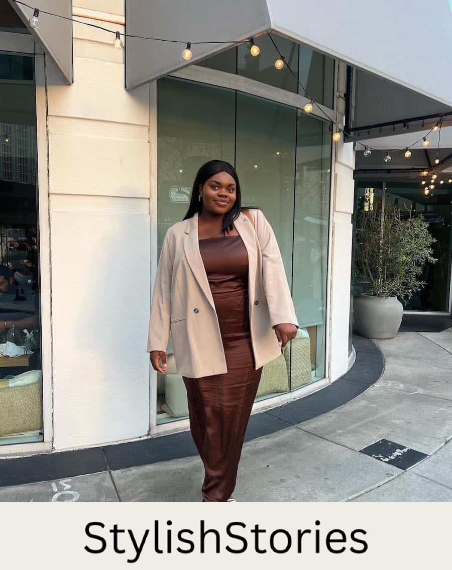 A woman wearing a silky brown maxi dress with a light beige blazer and strappy white heels