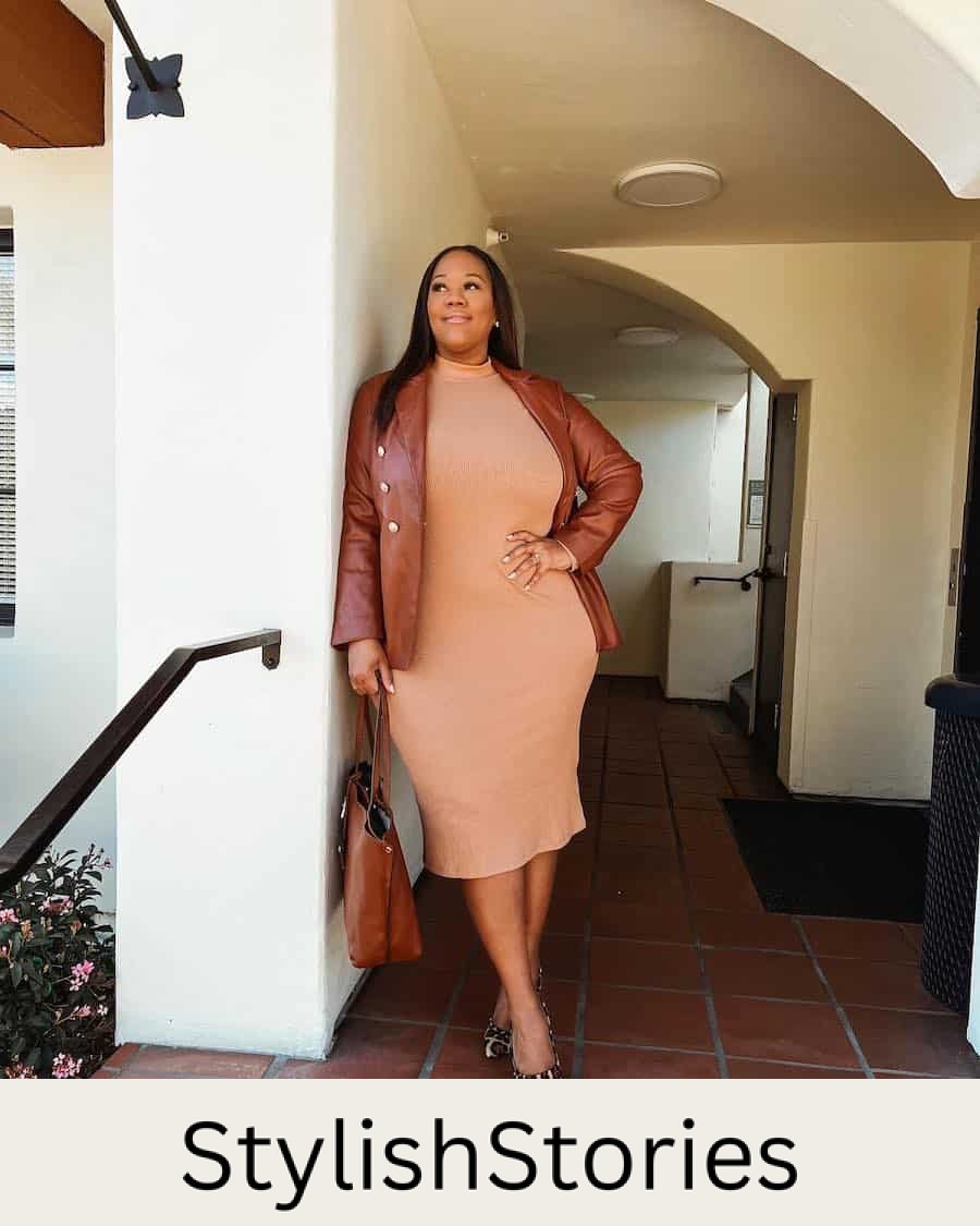 A woman wearing a mock neck camel midi dress with a brown leather jacket, a brown leather tote, and cheetah print ballet flats