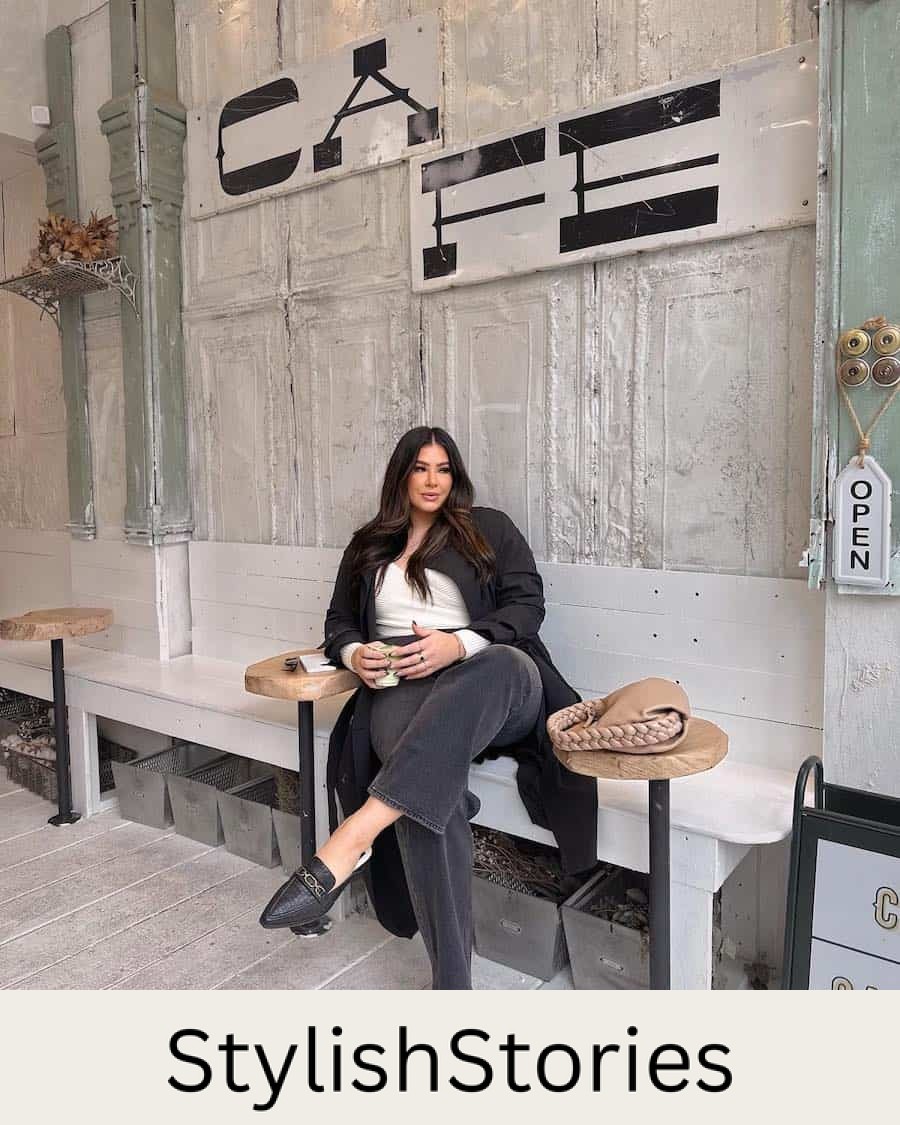 A woman wearing black jeans, a white tank top, a long black coat, and black loafers slides
