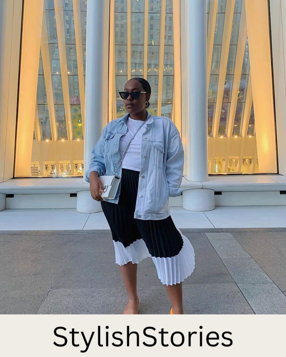 A woman wearing a black and white pleated midi skirt with a white top, a light blue denim jacket, and orange flats