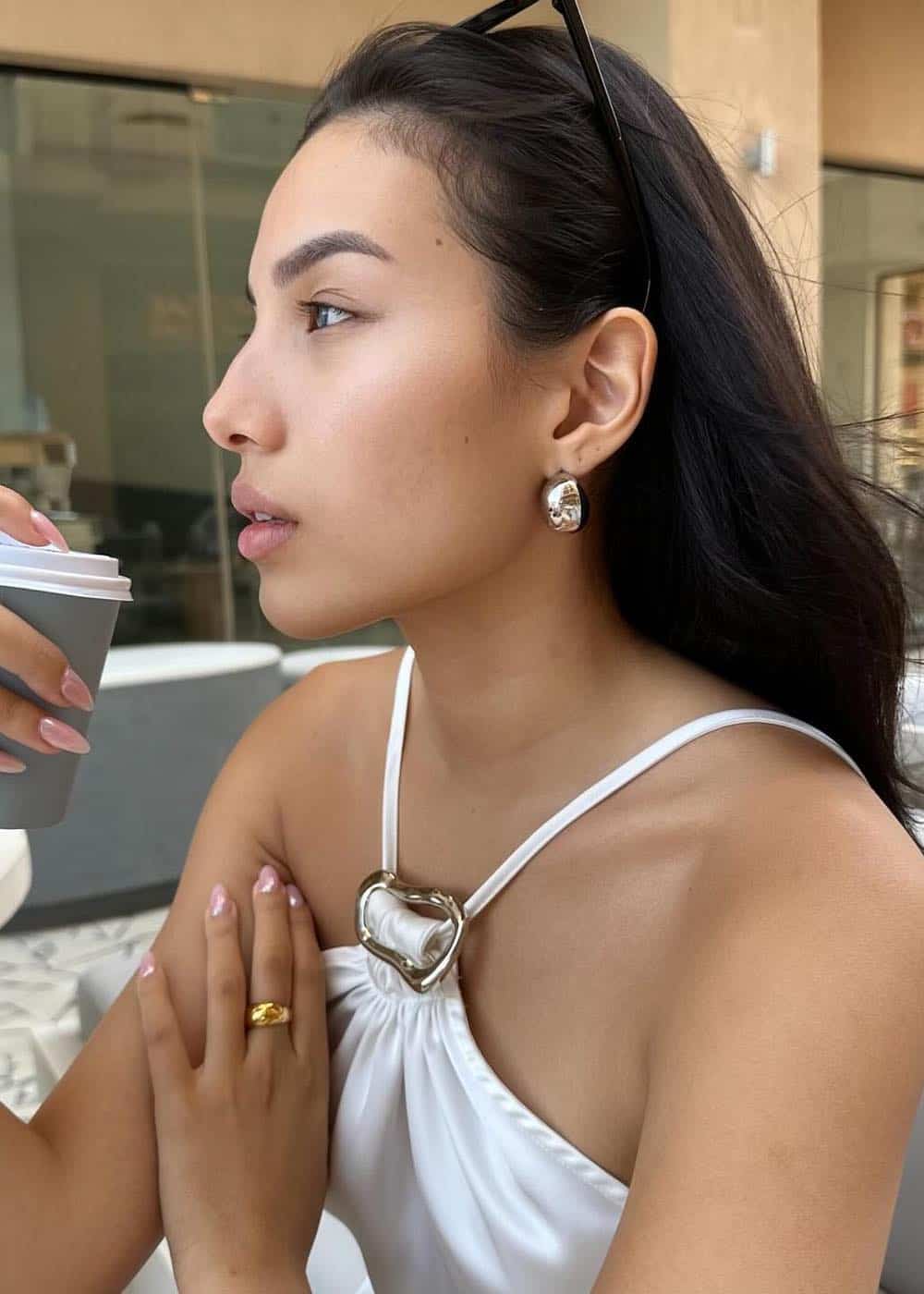 Close up shot of a woman drinking a to-go coffee while wearing a white skil halter top and gold jewelry.