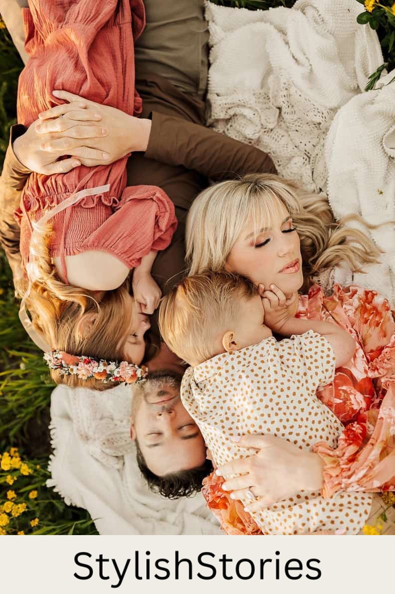 An image of a summer family photo with a family of four laying on a blanket in a field and holding each other, wearing red and pink toned clothing and neutrals