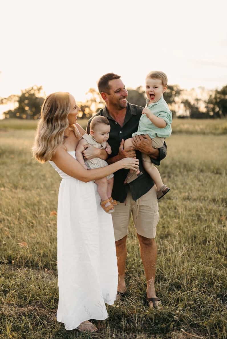 An image of a summer family portrait with a mother and father holding their two children, the mother wearing a white maxi dress and the other members wearing green and beige clothing