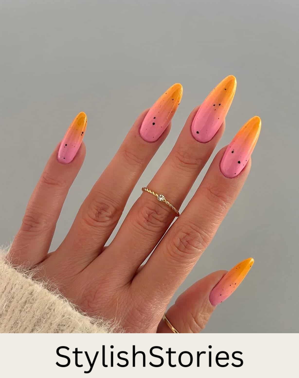 A hand with long almond nails featuring an orange and pink ombre with black speckles