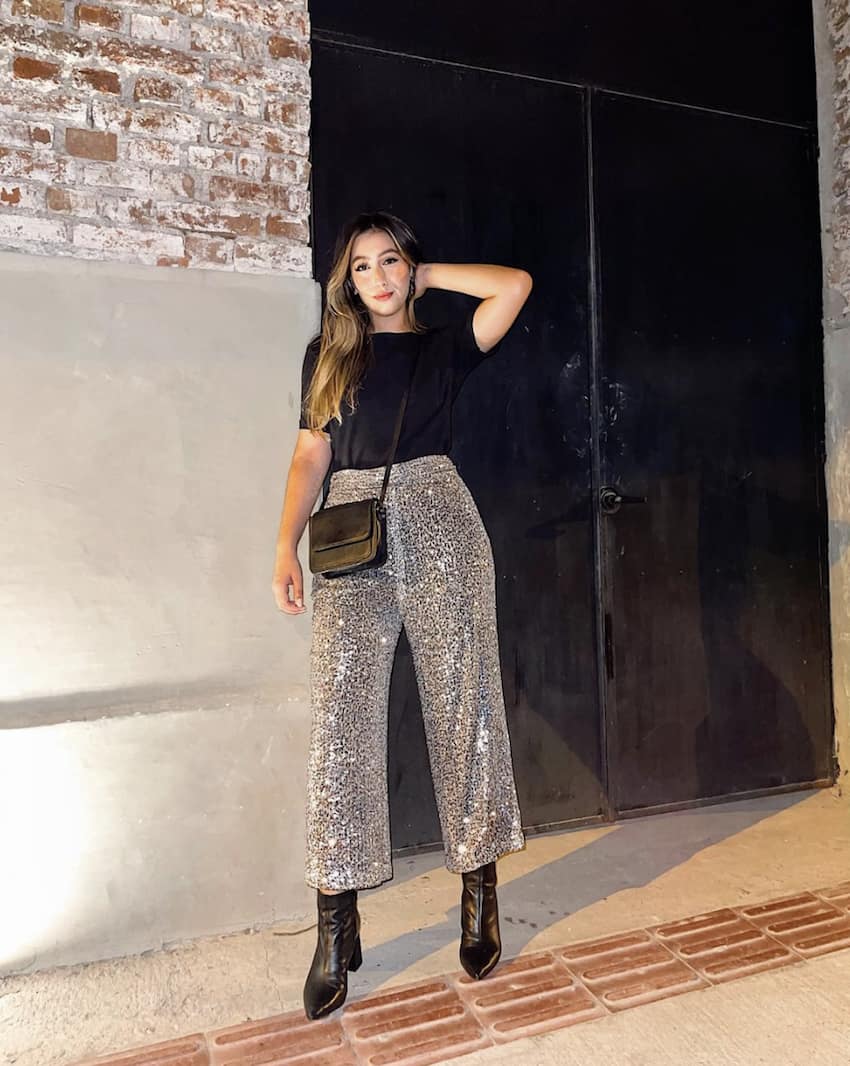 Image of a woman in a black t-shirt, silver sequin pants, black boots, and a black purse