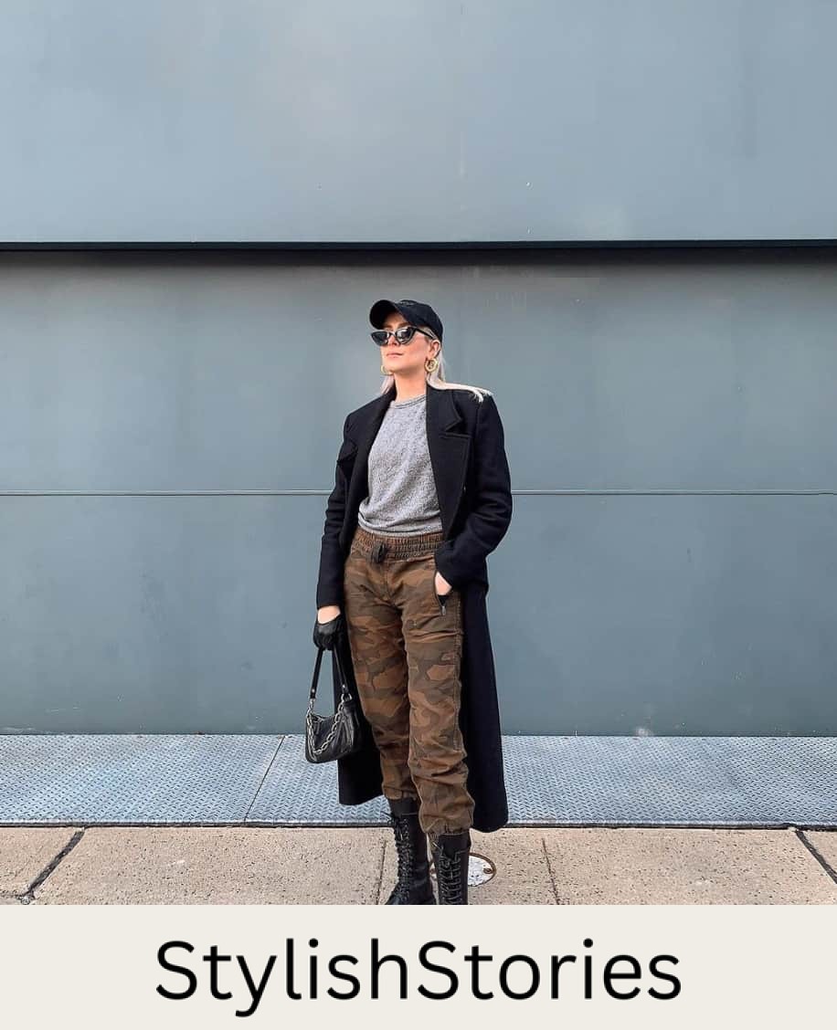 A woman wearing camo pants with combat boots, a grey top, and a long black coat and baseball cap
