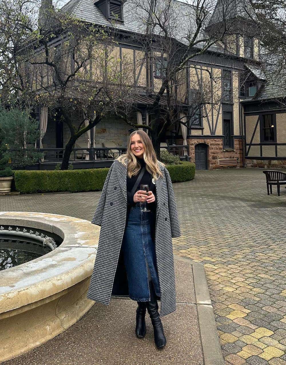 a woman wearing a denim midi skirt with a black sweater, a black and white plaid long coat, and black leather boots