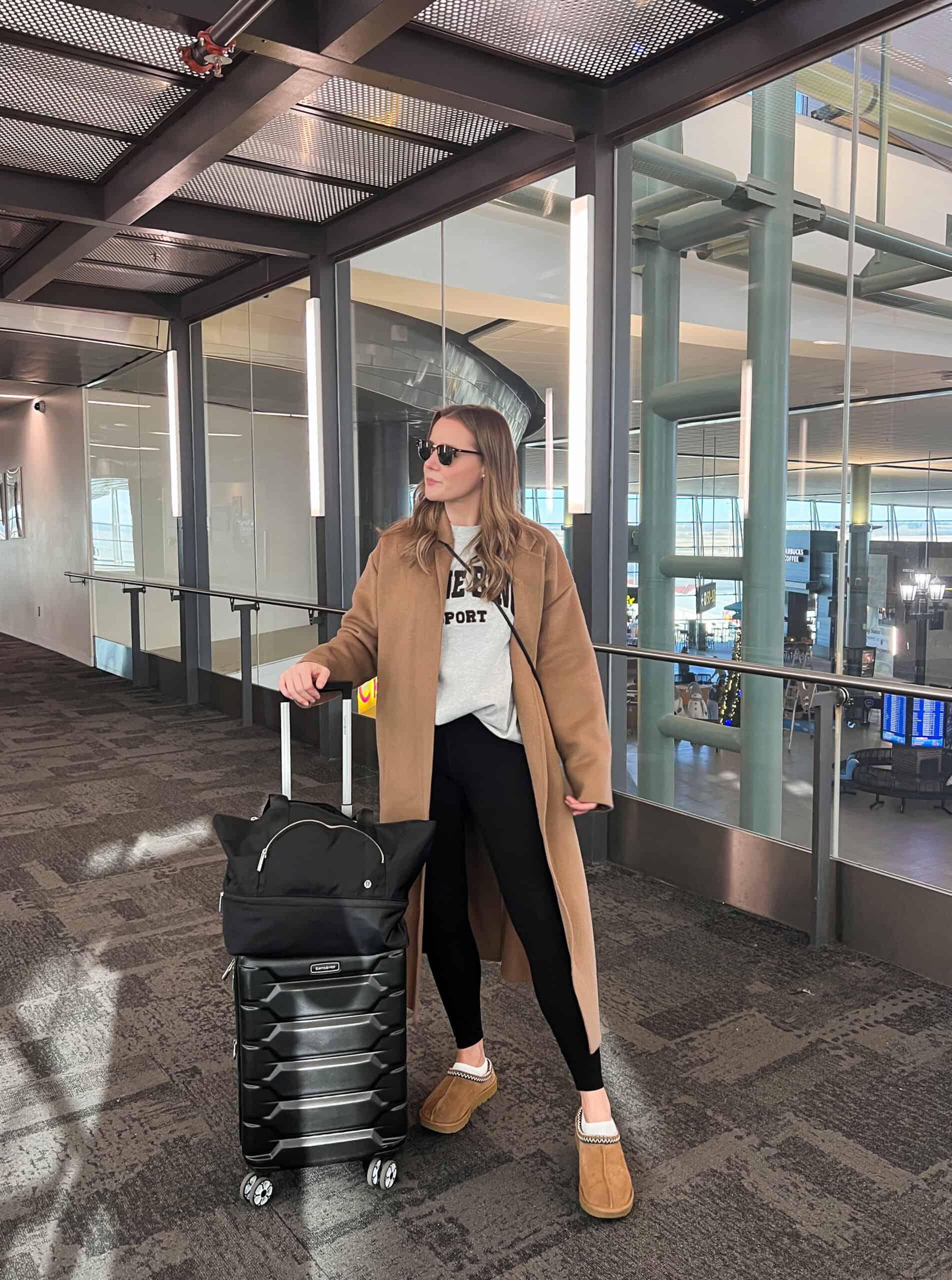 Christal wearing black leggings, a grey sweatshirt, a tan coat and Ugg tasman slippers in the airport.