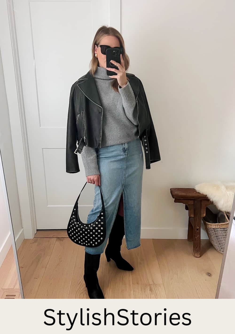 Christal wearing a long denim skirt with tall black boots, a grey sweater and a black leather jacket with a studded black bag.