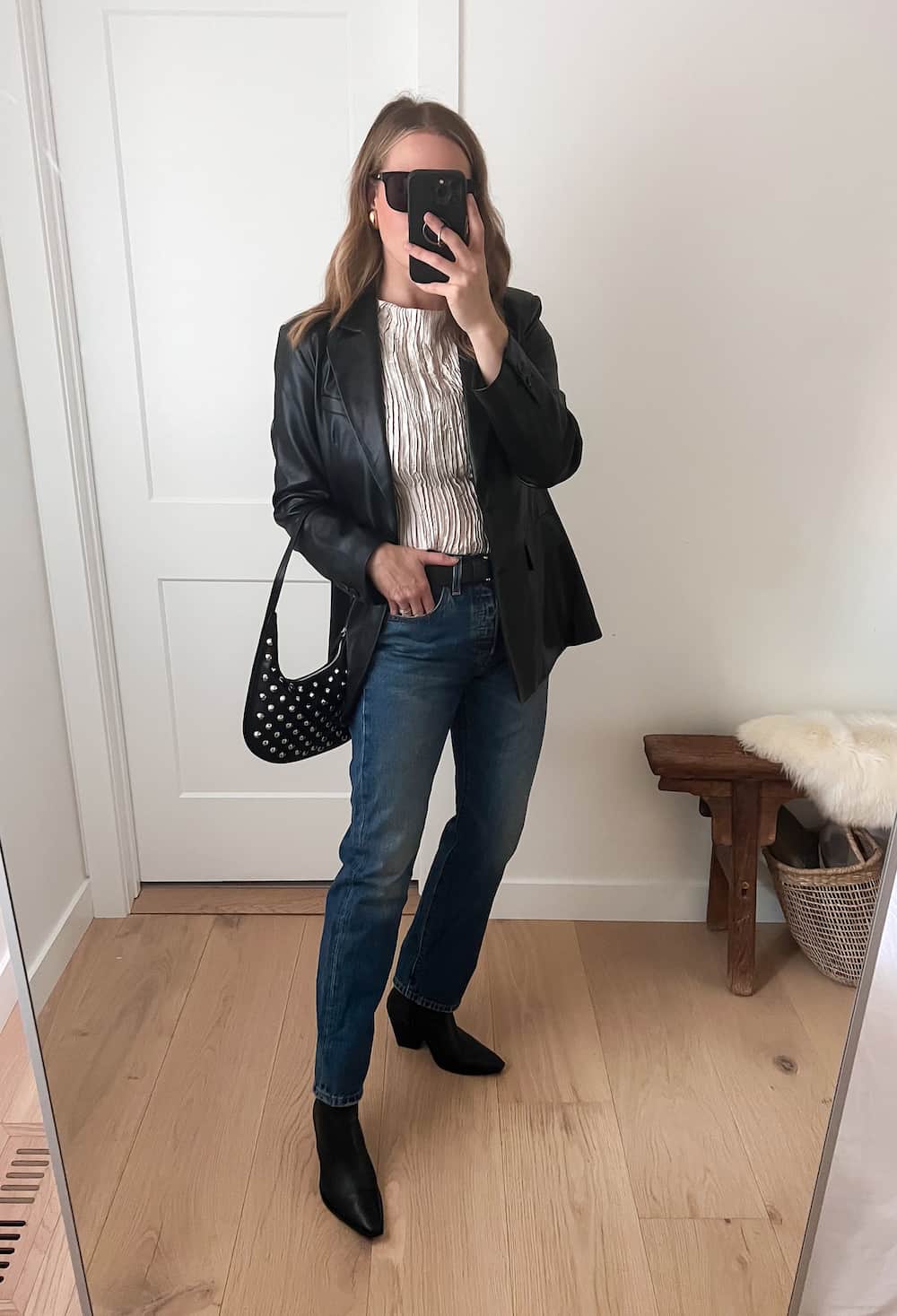 Woman wearing dark denim straight jeans, black booties, a silver top and a black leather jacket with a studded black bag.