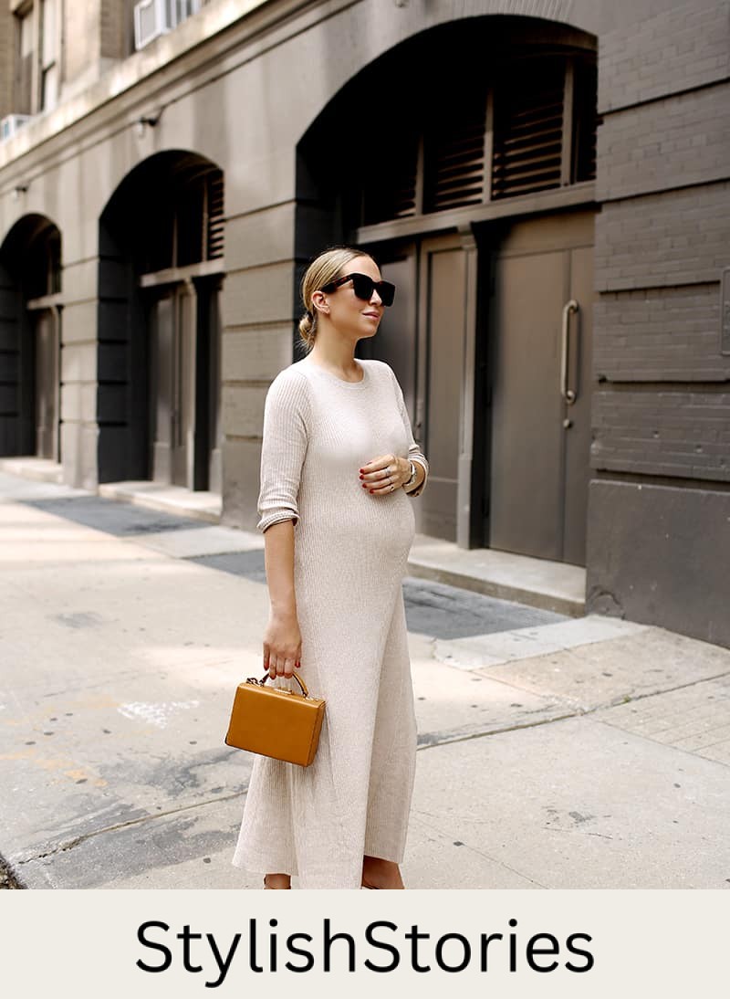 image of a pregnant woman standing on a sidewalk wearing a long cream colored sweater dress and dark sunglasses