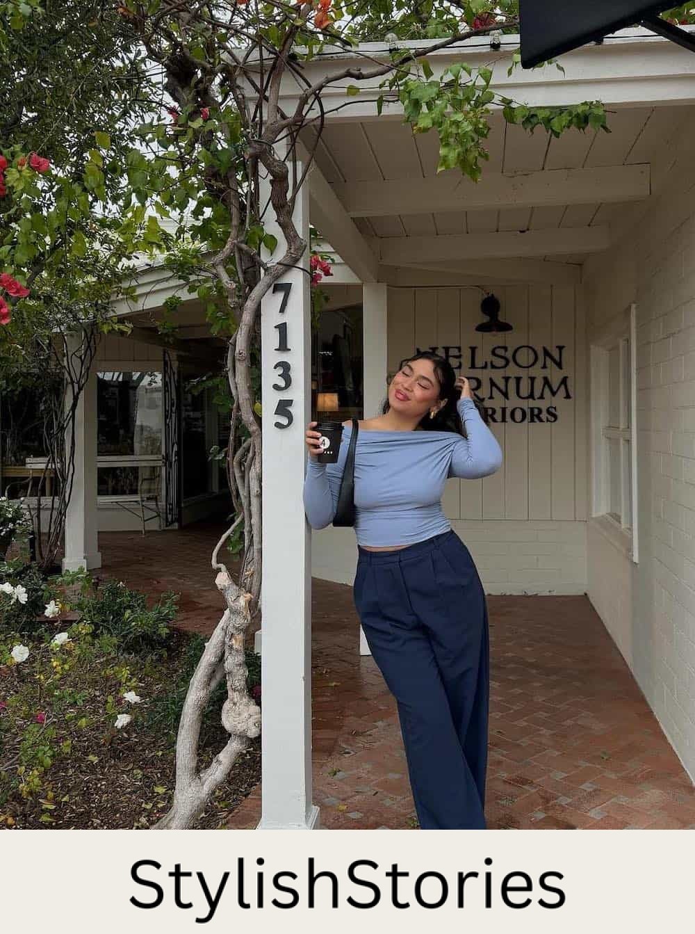 Woman wearing navy pants with a light blue off the shoulder top and sneakers.