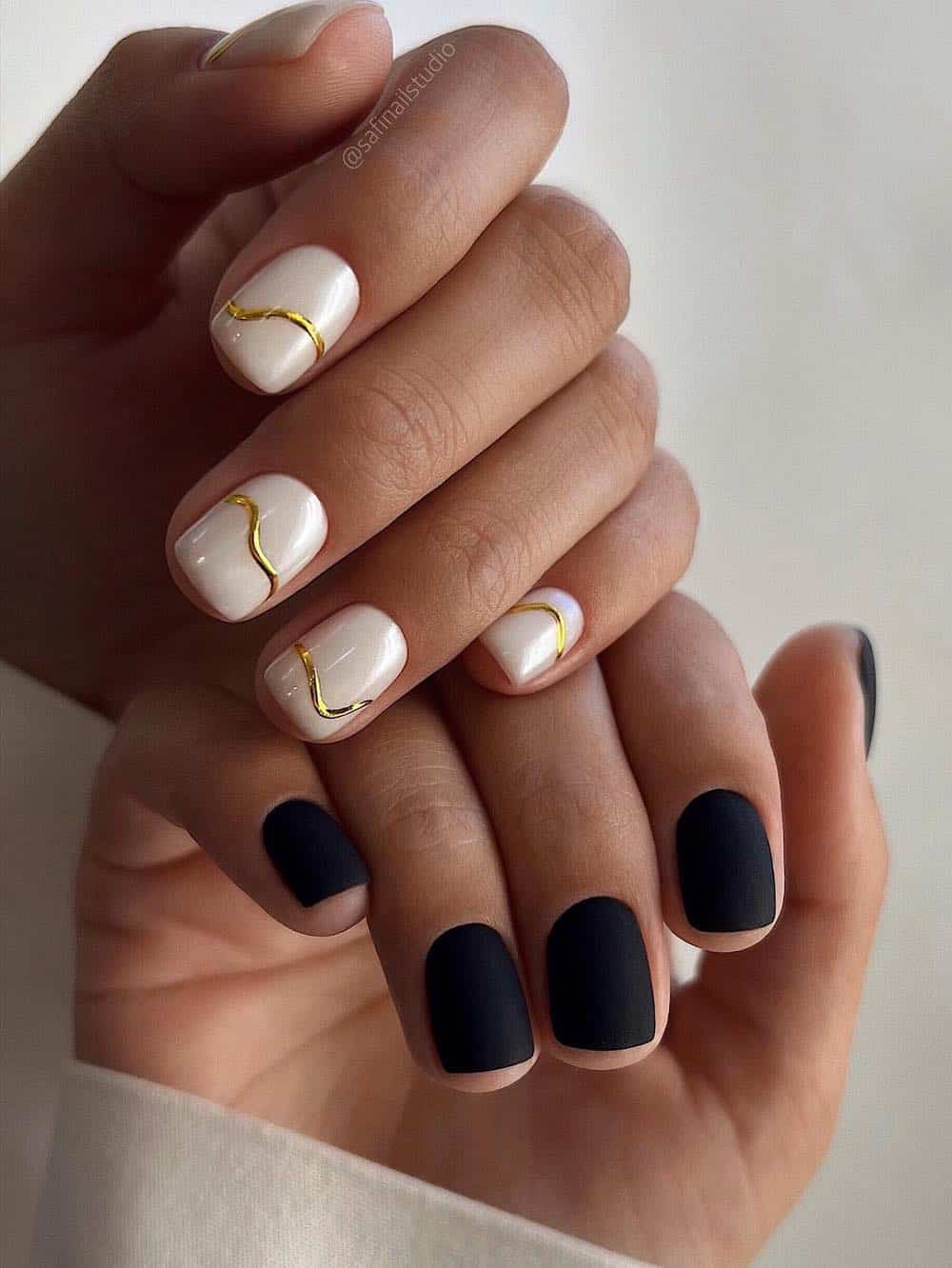a manicure featuring short matte black polish on one hand and glossy white polish with wavy gold accents on the other