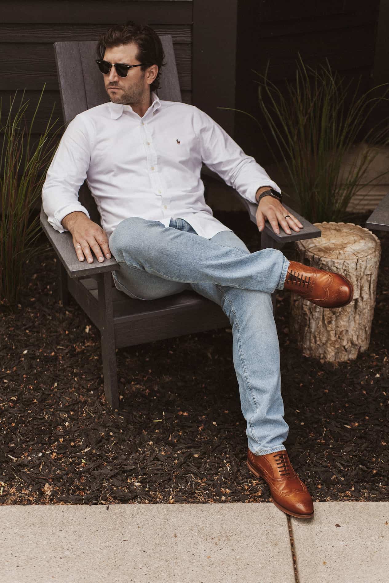 image of a man sitting on a black Adirondack chair wearing a white button-up shirt, light blue jeans, and brown dress shoes