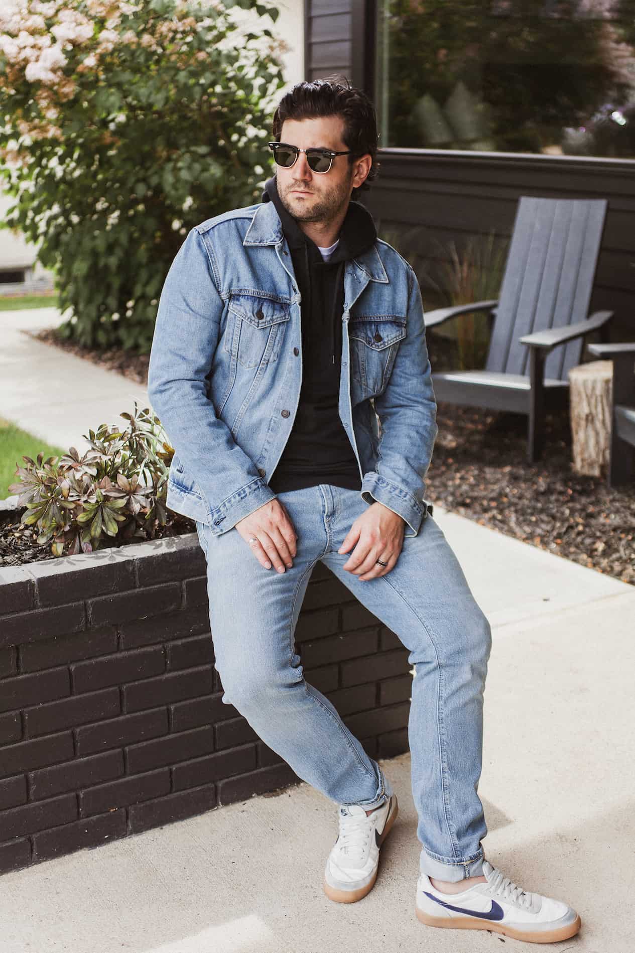 image of a man sitting on a brick planter wearing a denim jacket, black hoodie, light blue jeans, and white sneakers