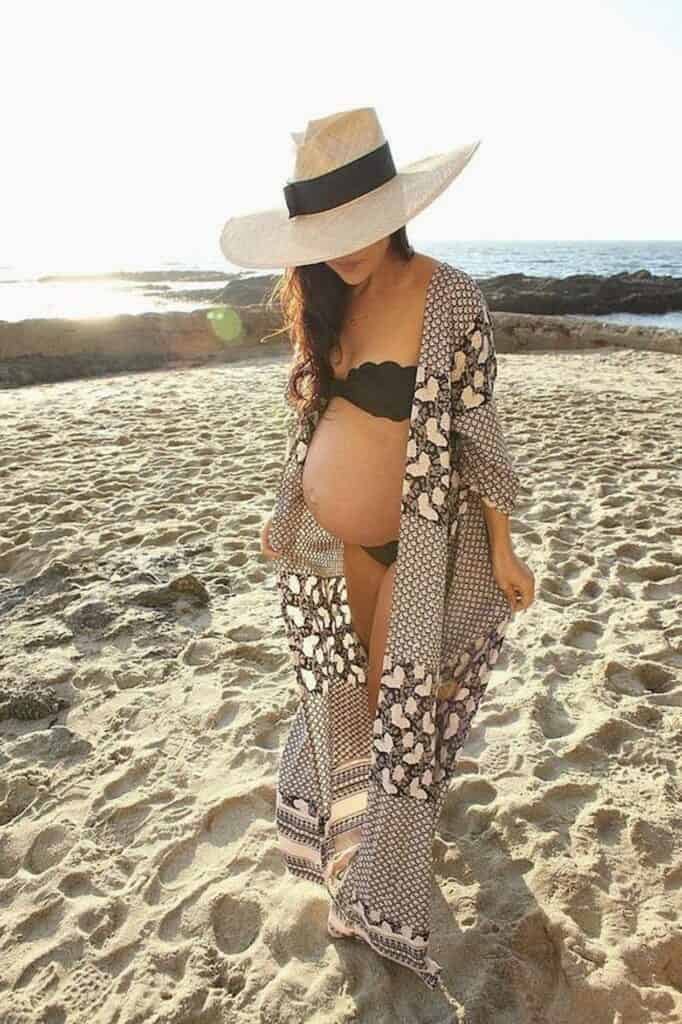 image of a pregnant woman walking on the beach wearing a large brim sun hat, black bikini, and a kimono robe over top