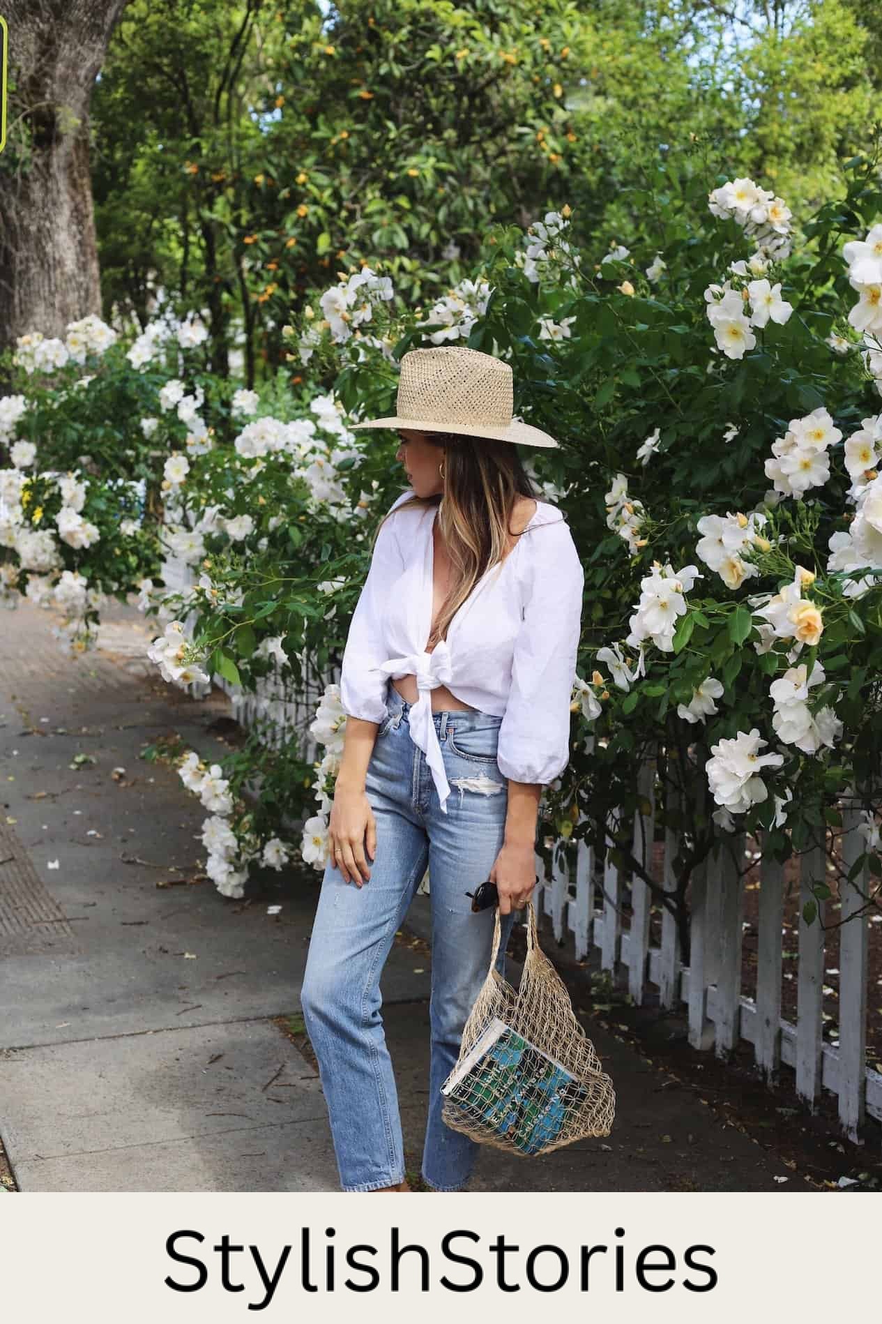 image of a woman standing on a sidewalk in front of flower bushes wearing a white blouse, blue jeans, and a woven hat