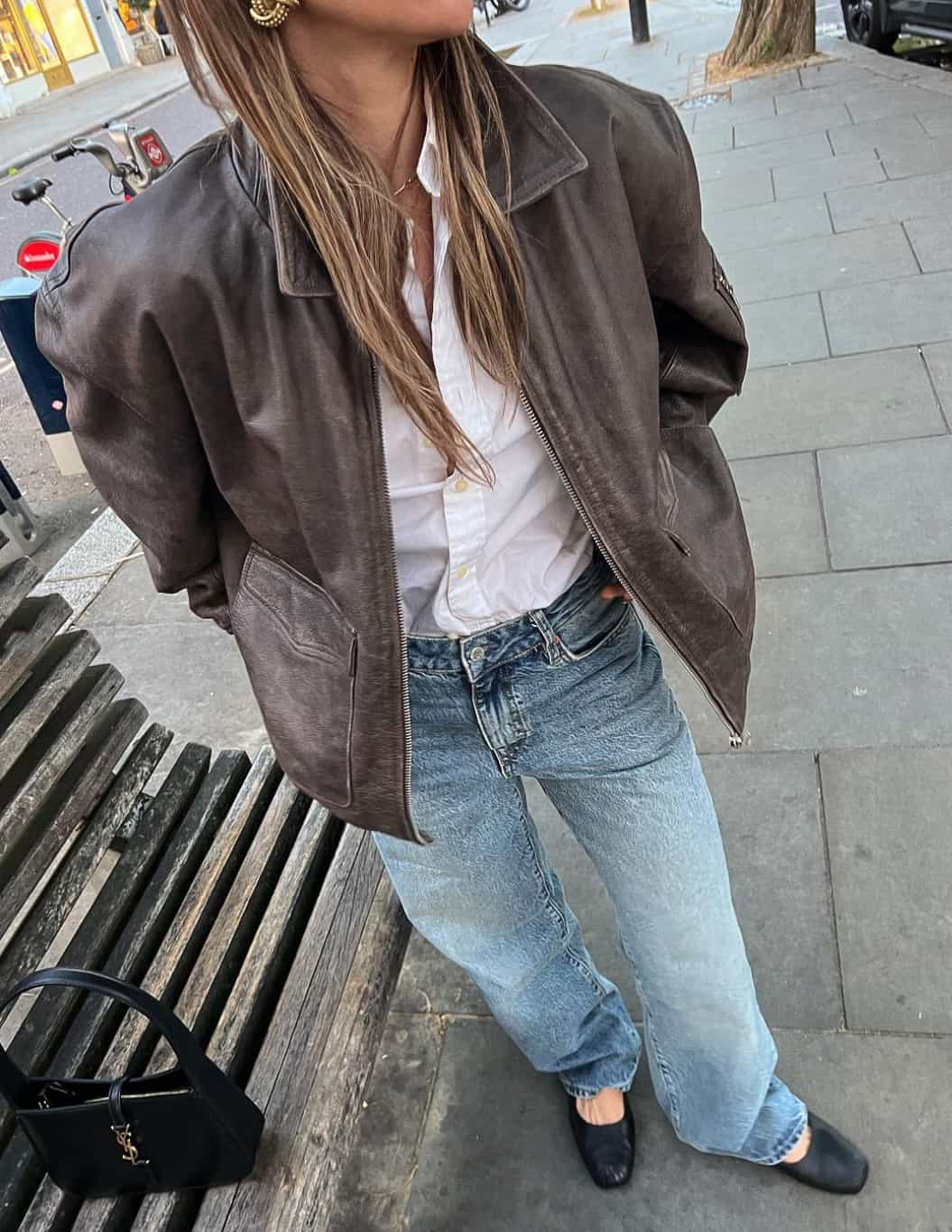 Woman wearing jeans, ballet flats, a white button down and a brown leather jacket.
